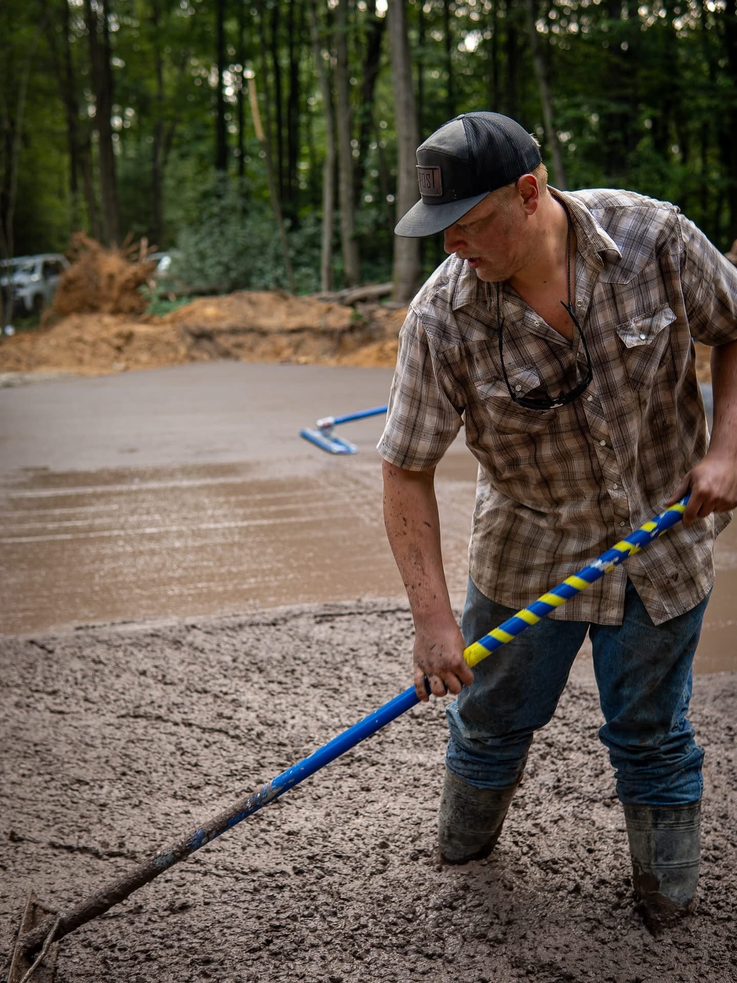 Man in a plaid shirt and cap levels wet concrete with a long-handled rake.