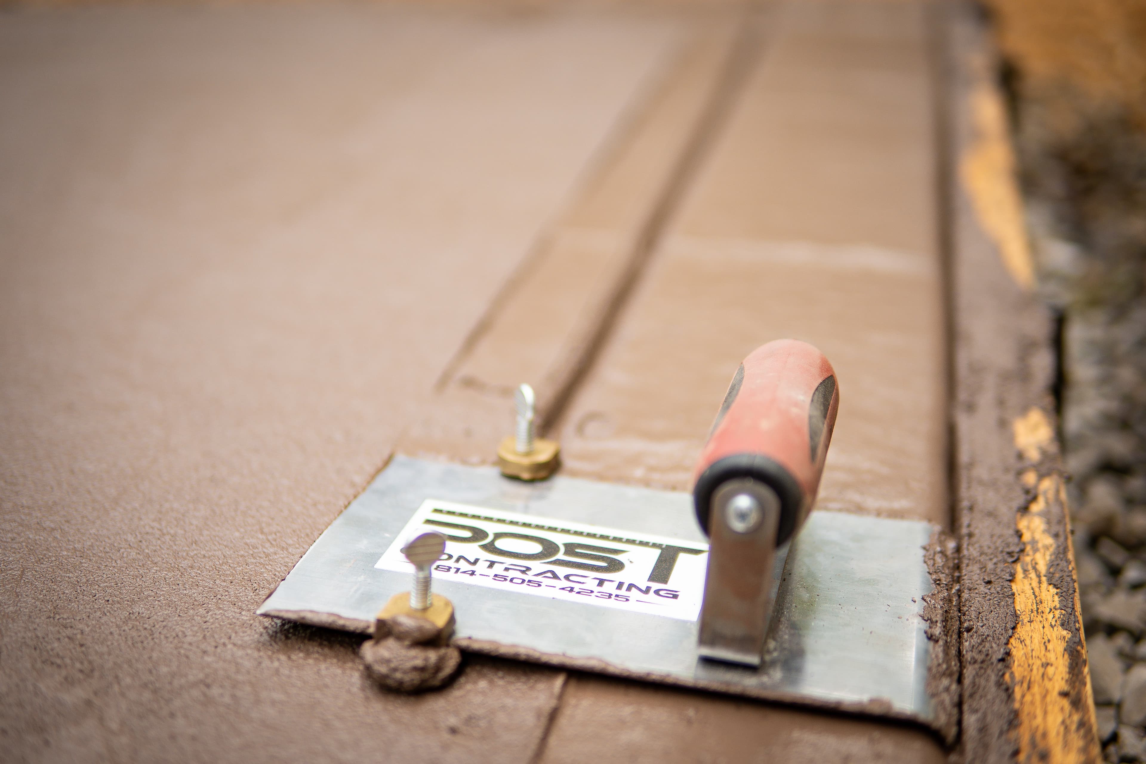 Post Contracting metal finishing tool resting on smooth, freshly poured brown-tinted wet concrete surface.