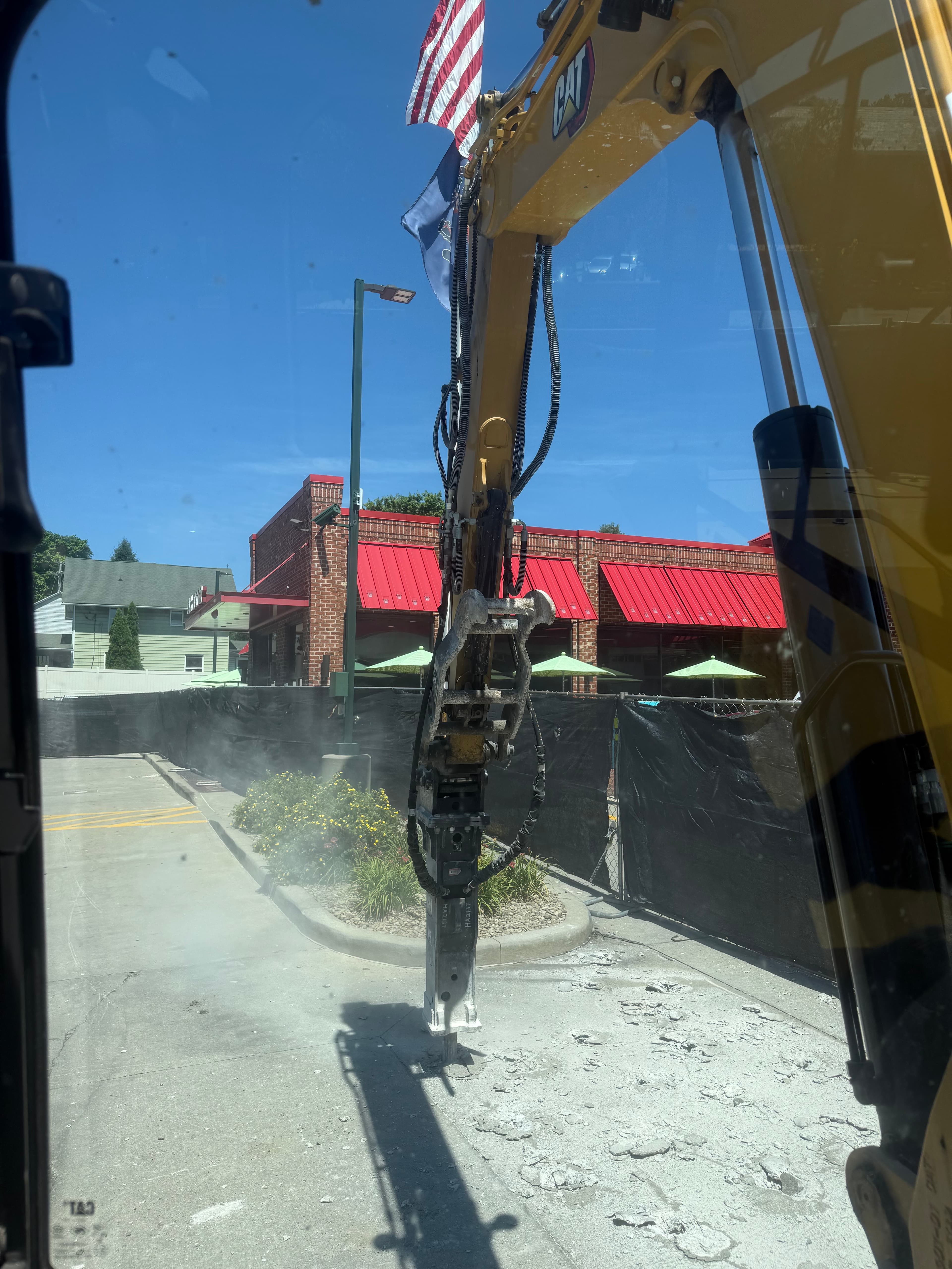 Excavator arm with a hydraulic hammer attachment working on concrete near a red building.