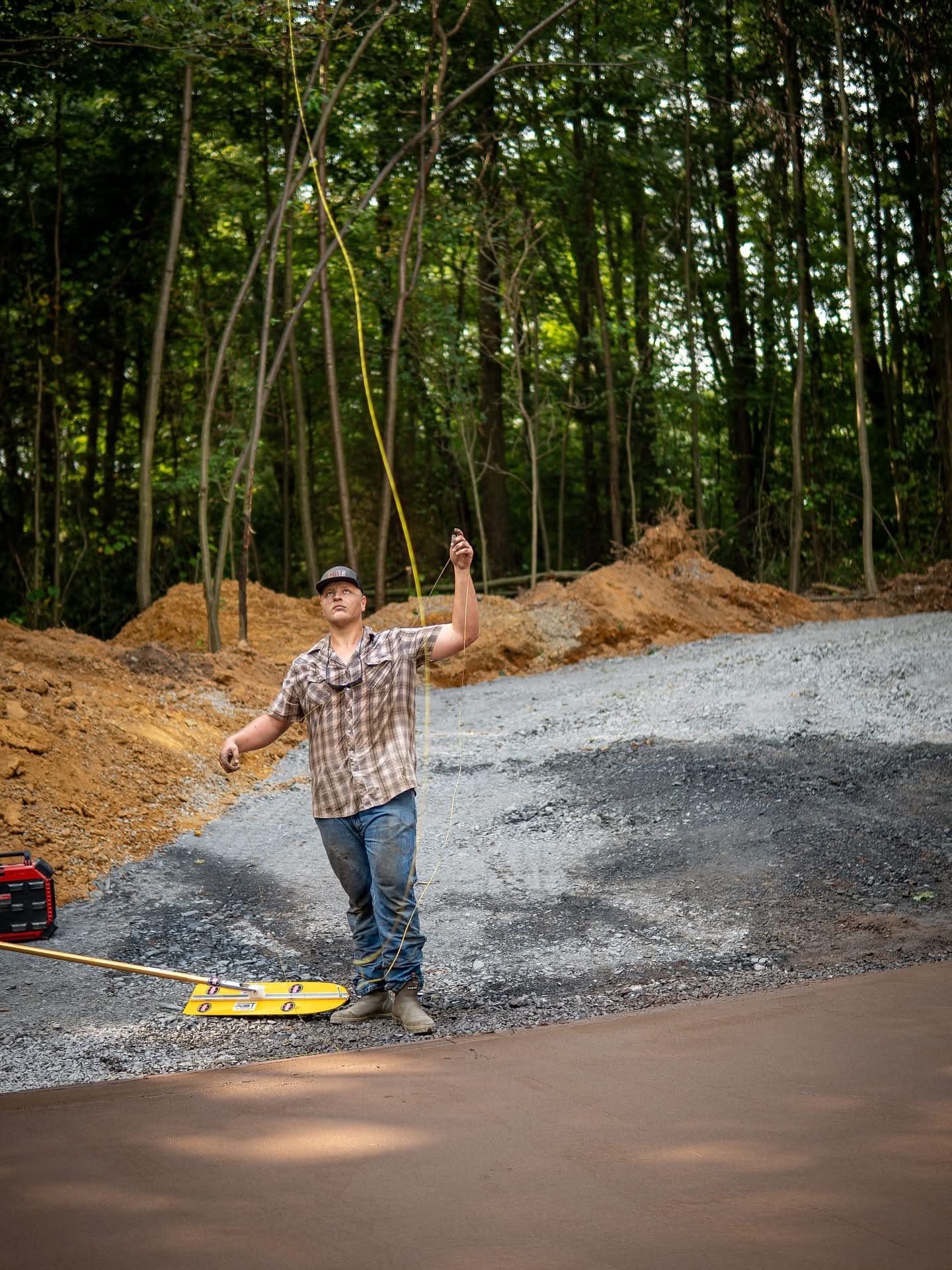 Man in plaid shirt holds a yellow line extending upwards at a wooded construction site.