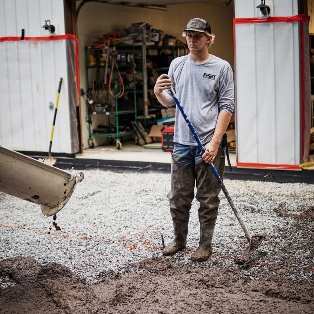 Construction worker in muddy waders holds a rake as concrete pours from a chute.